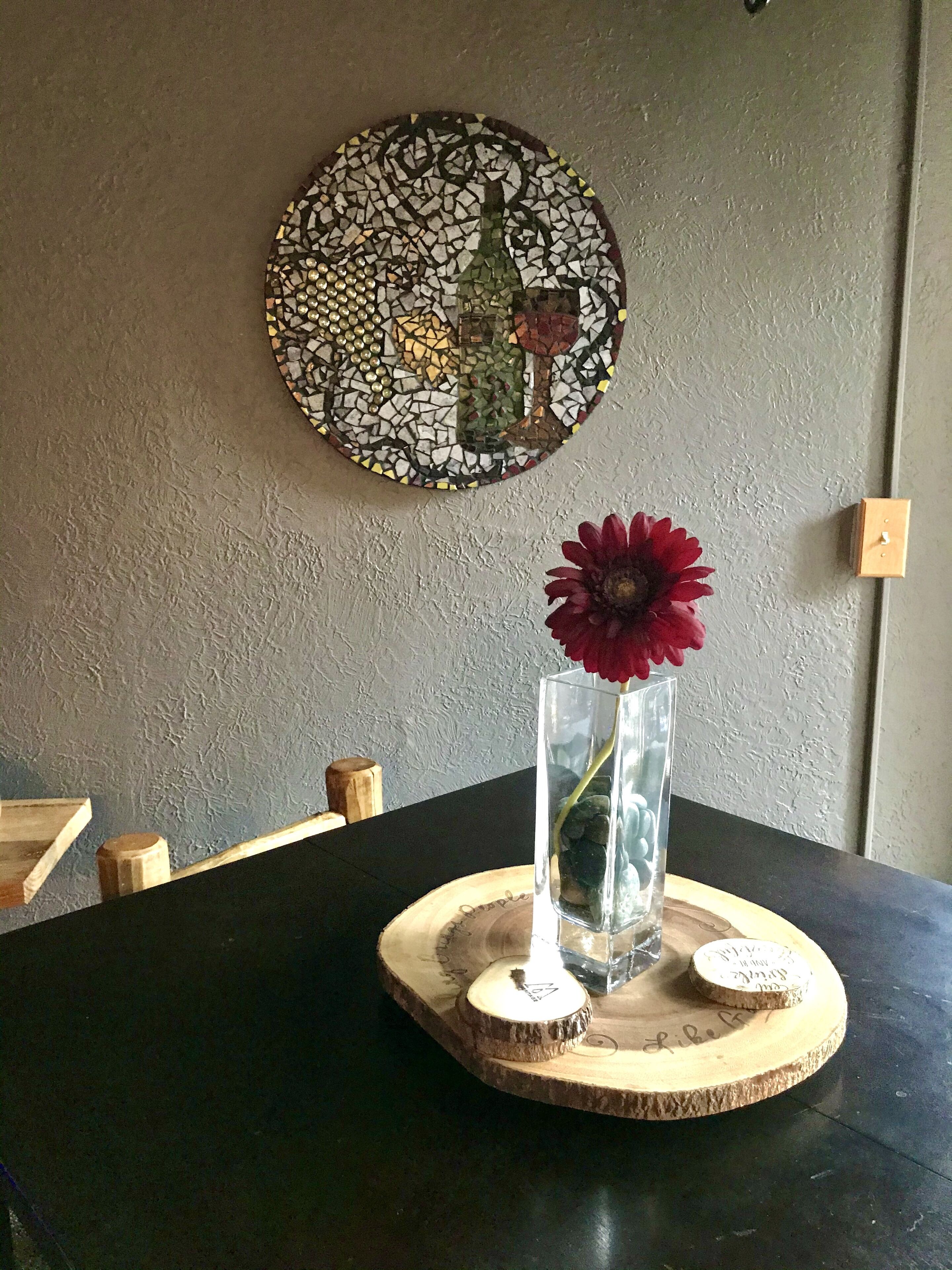 Une table à manger ornée d’un vase de fleurs rouges, une œuvre d’art en mosaïque accrochée au mur et un sous-verre en bois portant une inscription.