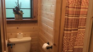 Et lille badeværelse i træ med toilet, et vindue med en plante og en bruseforhæng.