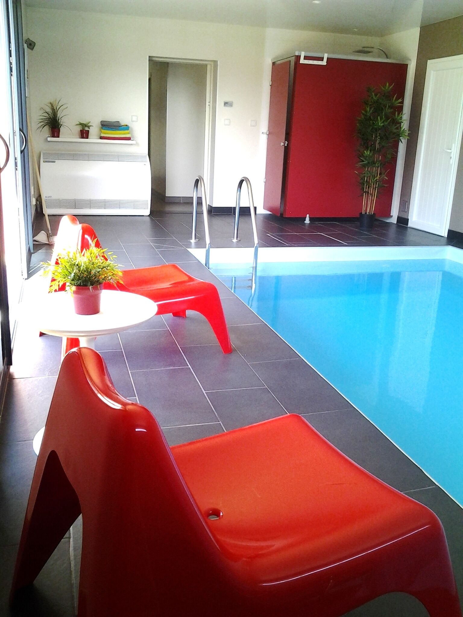 An indoor swimming pool with red chairs and a white table with a plant.