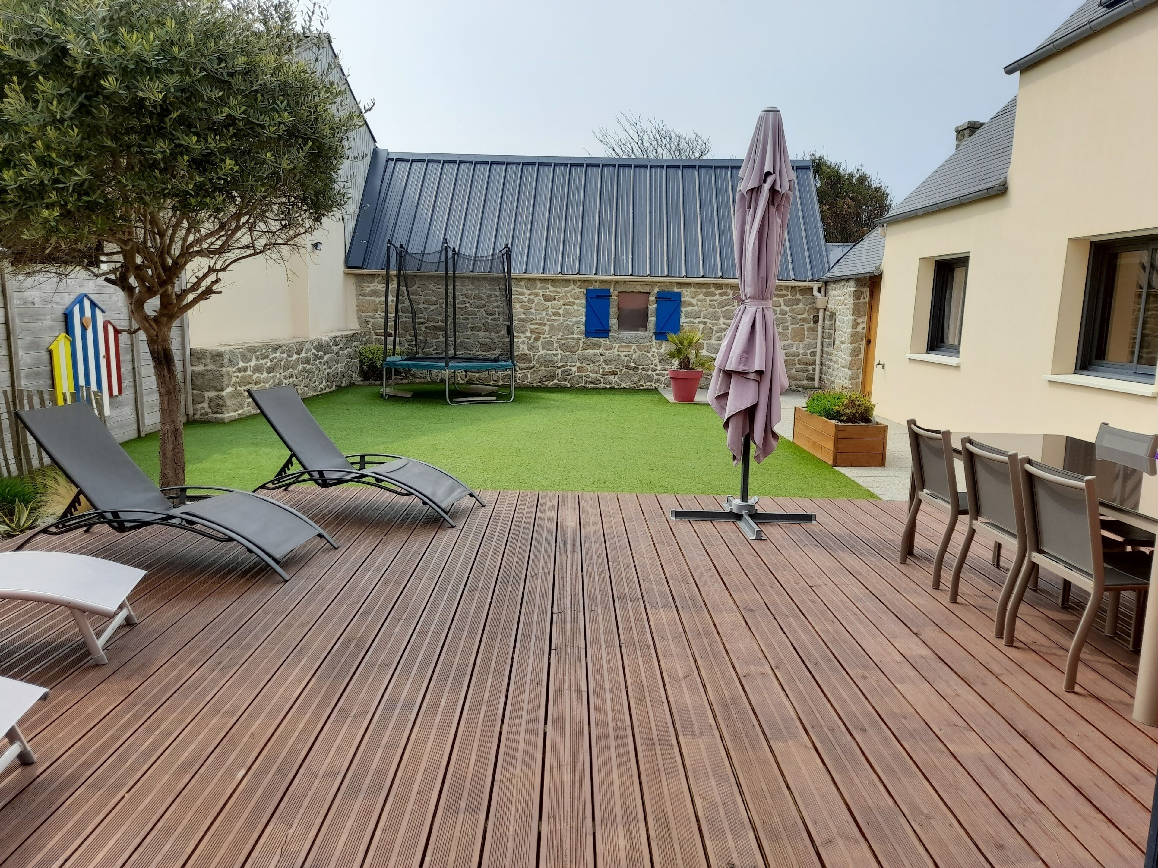 Una terraza de madera con sillones reclinables y una mesa, un muro de piedra y un edificio con techo azul al fondo.