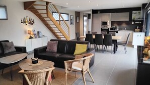 A modern living room with a sofa, coffee table, and wicker chairs. A staircase leads to a dining area with a table and chairs. The kitchen is visible with cabinets and appliances.