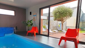 An indoor swimming pool with clear blue water, surrounded by red chairs and a large window offering a view of the outside.