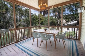 A covered outdoor area with a wooden table and chairs, surrounded by glass walls offering views of trees and a building.