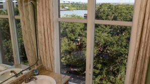 A bathroom with a sink, mirror, and window overlooking a green landscape with a bridge in the distance. - Step into a spacious, luxury condo that has it all – a premiere location in the heart of the Historic District and floor-to-ceiling window views of the Savannah River, Talmadge Bridge and City Hall. 5th floor.Best river views in the building. (Savannah)