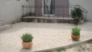 A courtyard with gravel, two potted plants, a metal railing, and a building with a door and windows. - Charming studio, quiet, patio, 2-4p (Fontvieille)
