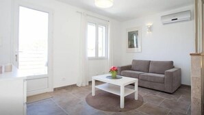 A bright, white room with a grey sofa, a small white table with a vase, and a large window. - Charming studio, quiet, patio, 2-4p (Fontvieille)