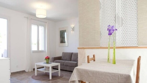 A bright living room with a white sofa, a small white table with a vase of flowers, and a dining table with chairs. - Charming studio, quiet, patio, 2-4p (Fontvieille)