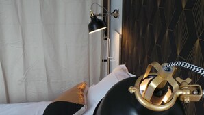 Ein ordentlich bezogenes Bett mit weißen und goldenen Kissen, einer schwarzen und goldenen Lampe sowie einer Wand mit geometrischen Mustern.