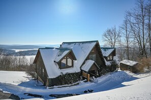 Une cabane dotée d’un toit en pente, entourée de terre et d’arbres recouverts de neige, offrant une vue dégagée sur le paysage en arrière-plan.