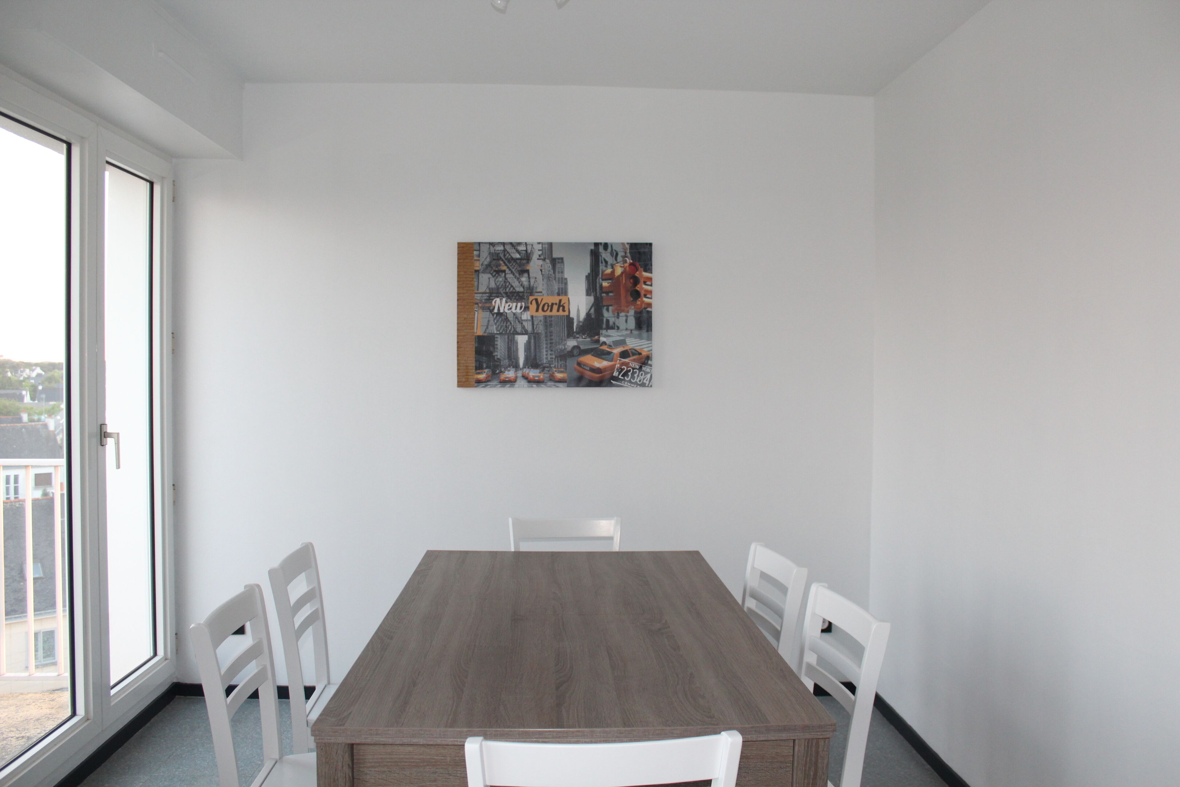 Uma sala de jantar com mesa de madeira e cadeiras brancas, um quadro com vista urbana na parede e uma janela grande à esquerda.