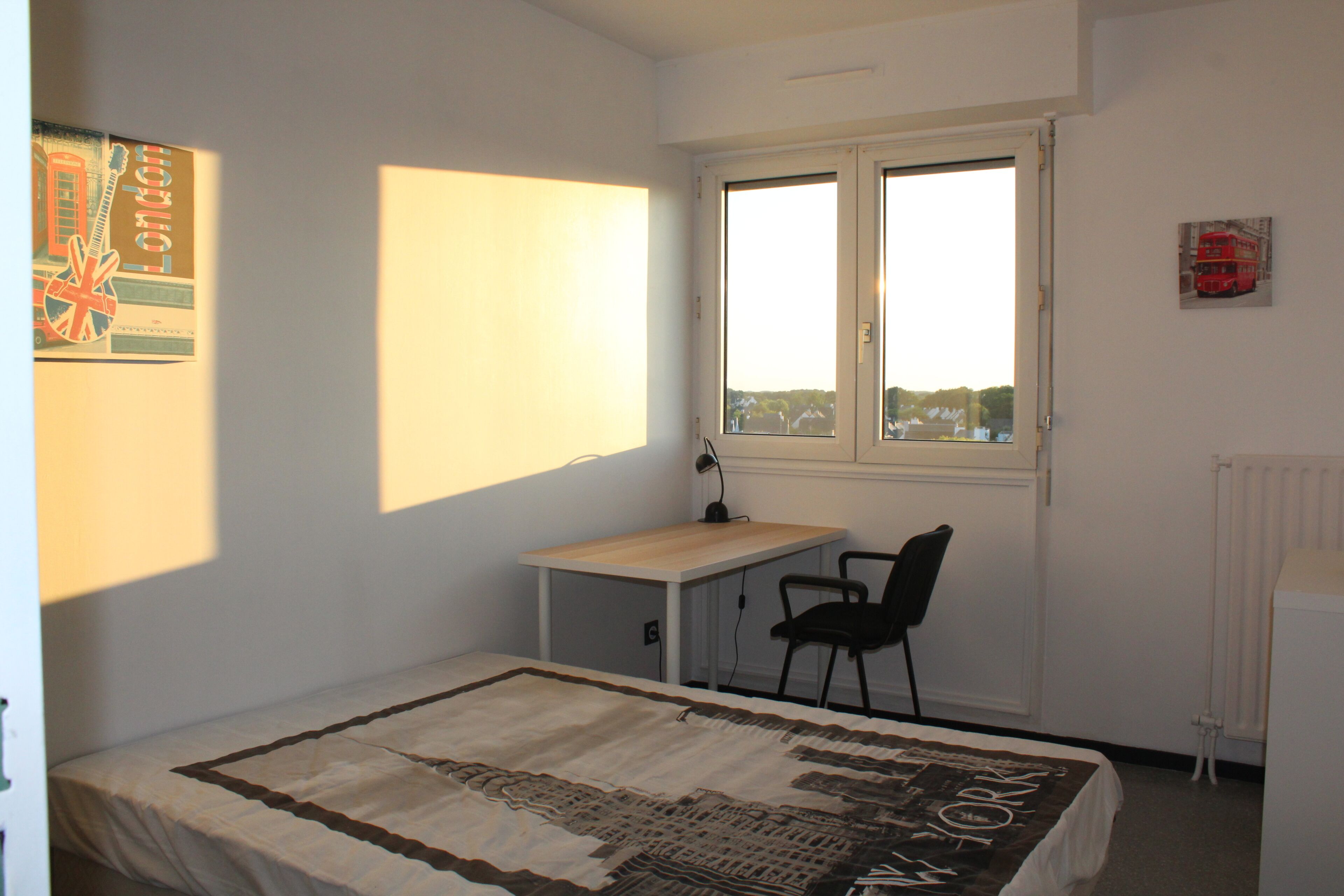 Une chambre avec un lit simple, un bureau et une chaise. Il y a une fenêtre donnant sur les bâtiments environnants.