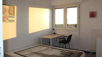 Une chambre avec un lit simple, un bureau et une chaise. Il y a une fenêtre donnant sur les bâtiments environnants.