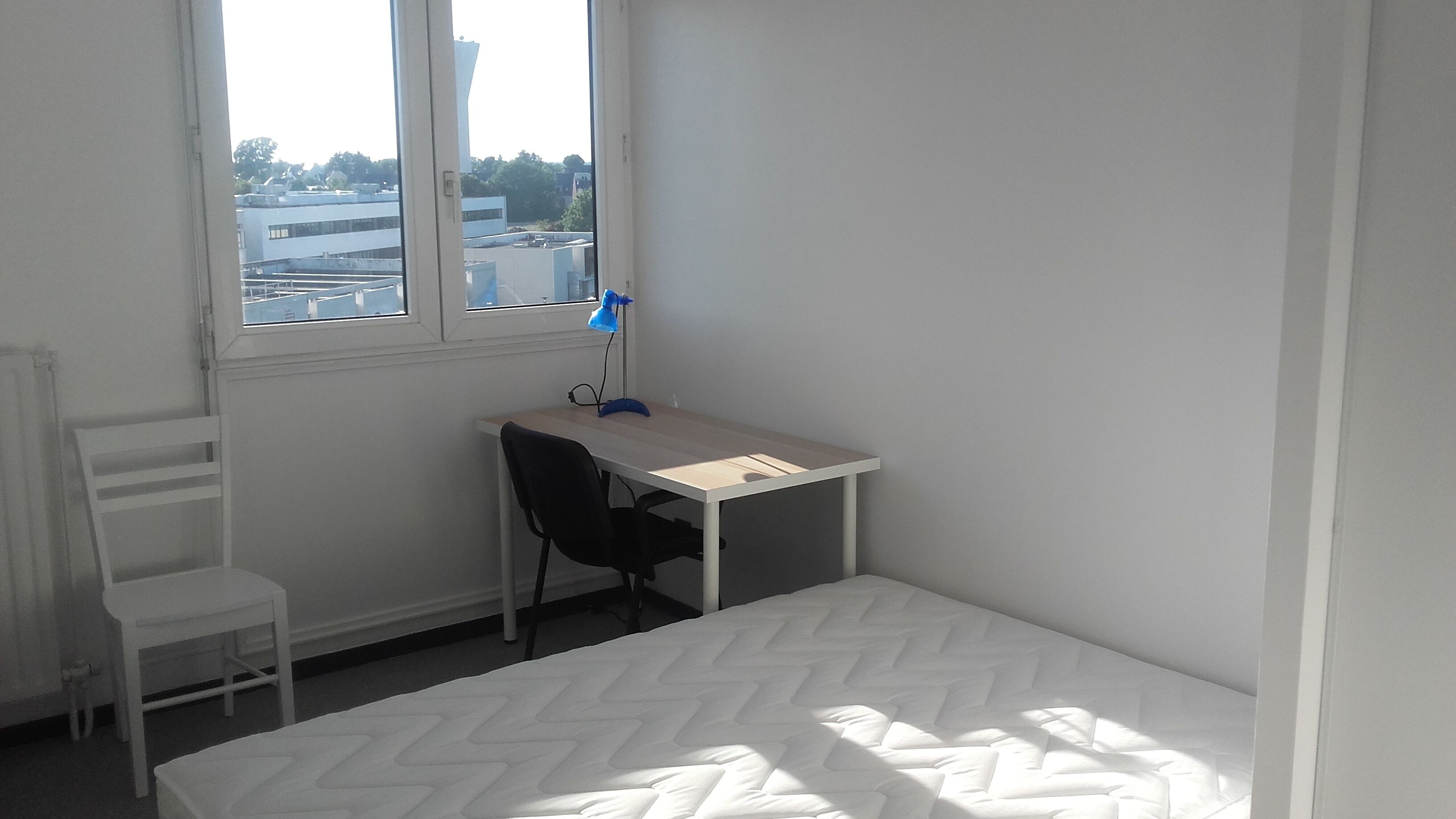 A small, white room with a single bed, a desk with a chair, and a window.