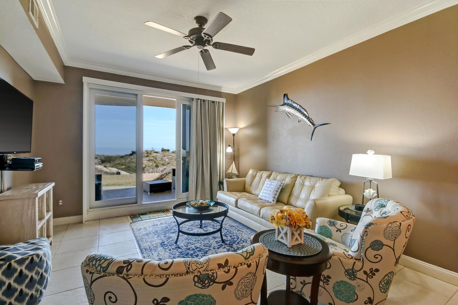 A spacious living room with a large sliding glass door leading to a balcony, a ceiling fan, a sofa with patterned upholstery, and a small round table with a decorative centerpiece.