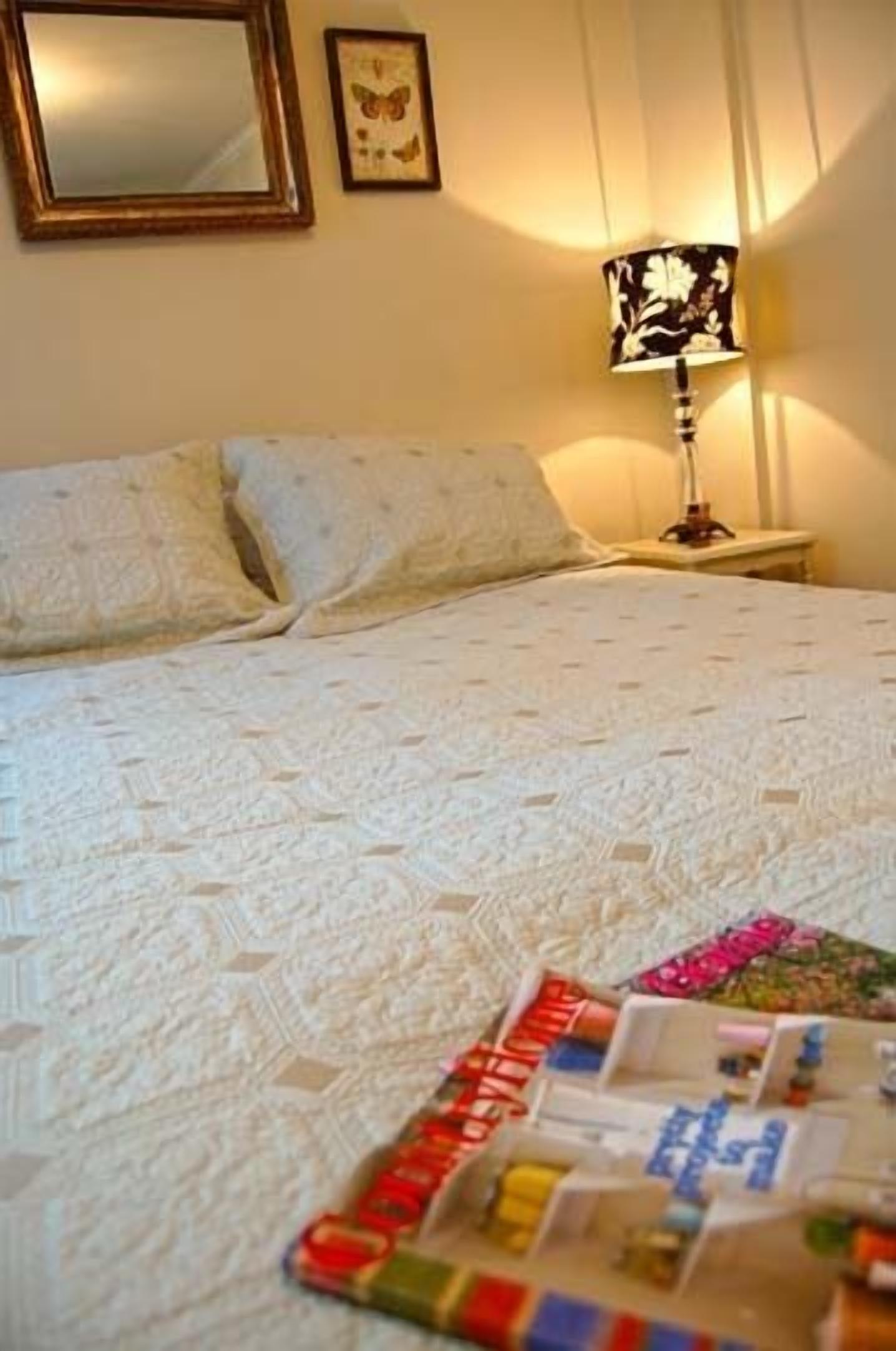 Een net opgemaakt bed met een bedsprei met een patroon, een bedlampje en een lijst met een afbeelding van een vlinder aan de muur.