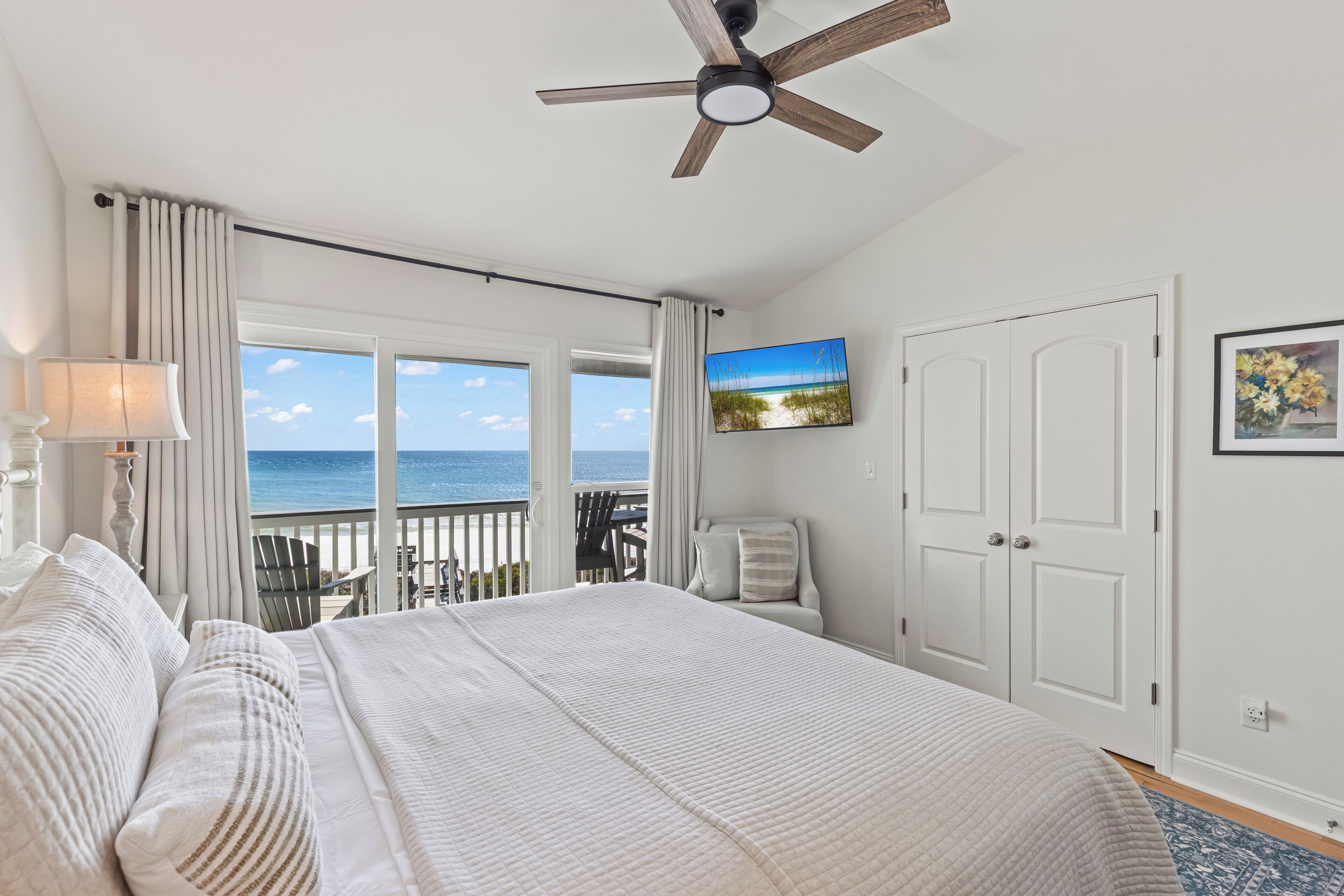 A bedroom with a large bed, a ceiling fan, a view of the ocean, a TV on the wall, and a door.