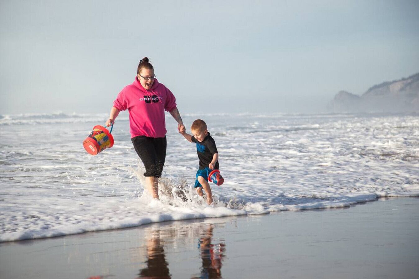 Eine Person in einem rosafarbenen Hemd und schwarzer Hose geht am Strand entlang, ein Kind dabei, das einen roten Eimer hält.