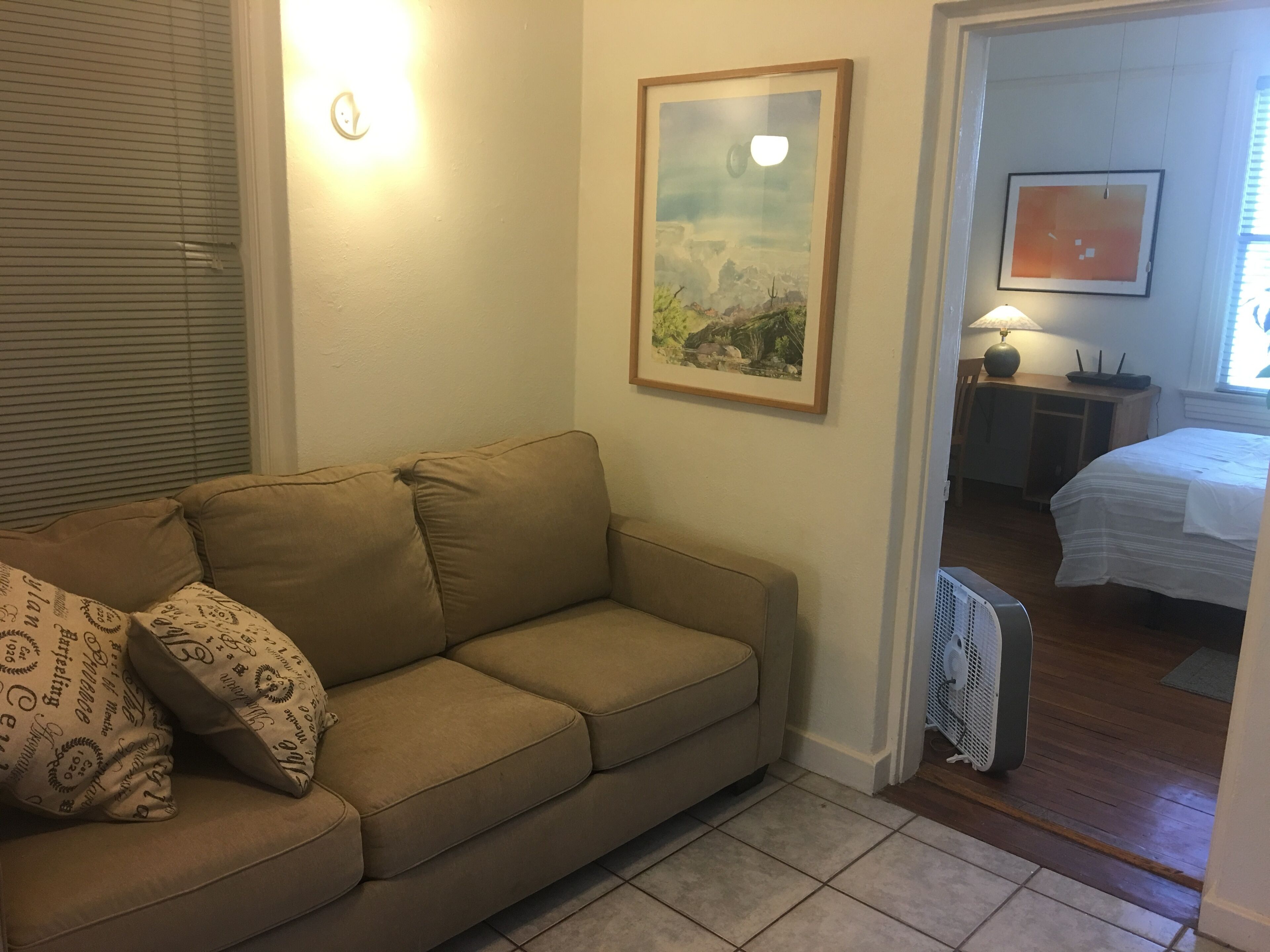 A living room with a beige sofa, a framed landscape painting, a bedside table with a lamp, and a bed with a white bedspread.