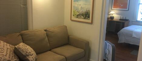 A living room with a beige sofa, a framed landscape painting, a bedside table with a lamp, and a bed with a white bedspread.