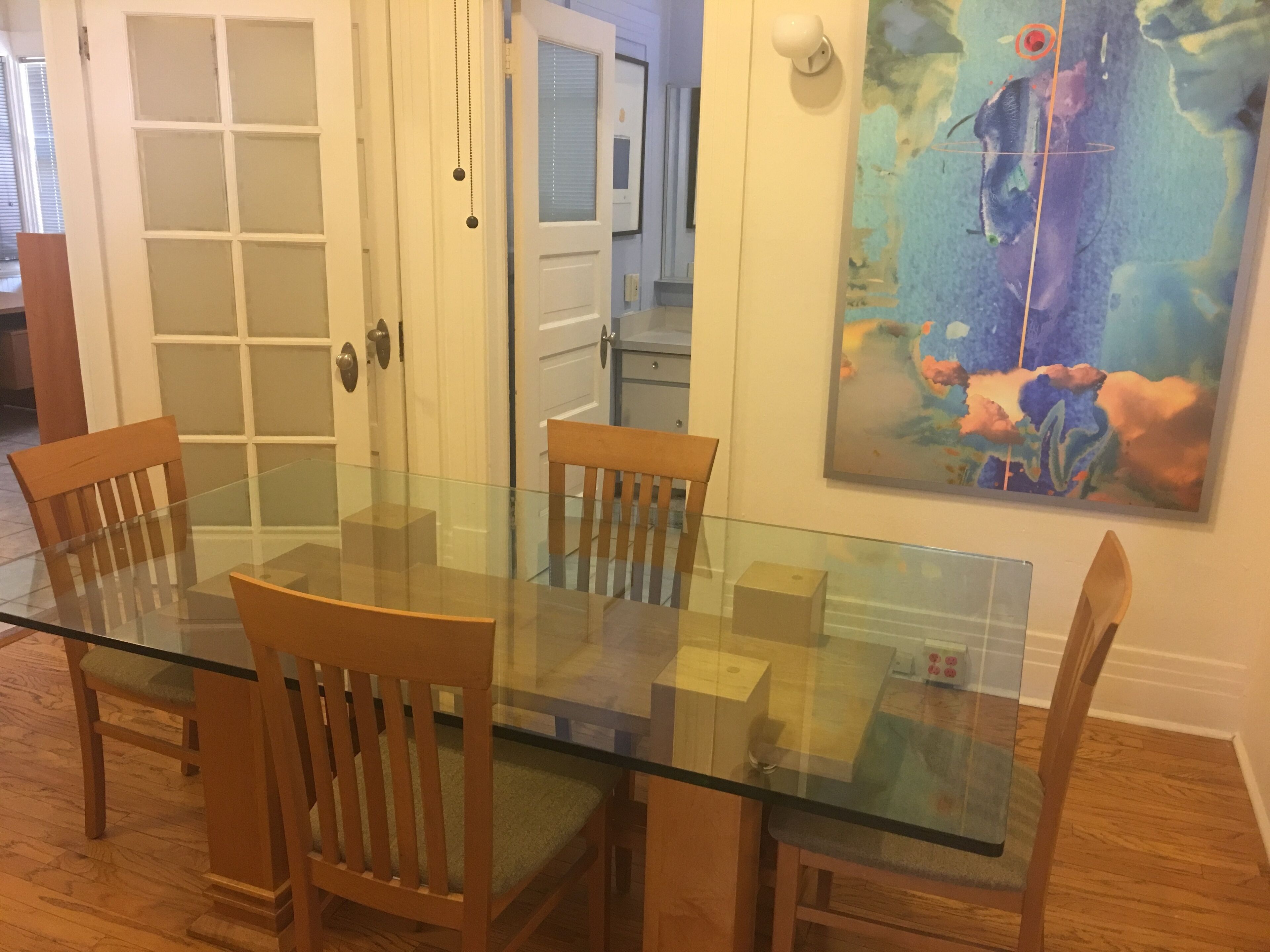 Ein Essbereich mit einem Glastisch und Holzkästen, eine weiße Tür mit satinierten Glasscheiben und ein farbenfrohes abstraktes Gemälde an der Wand.