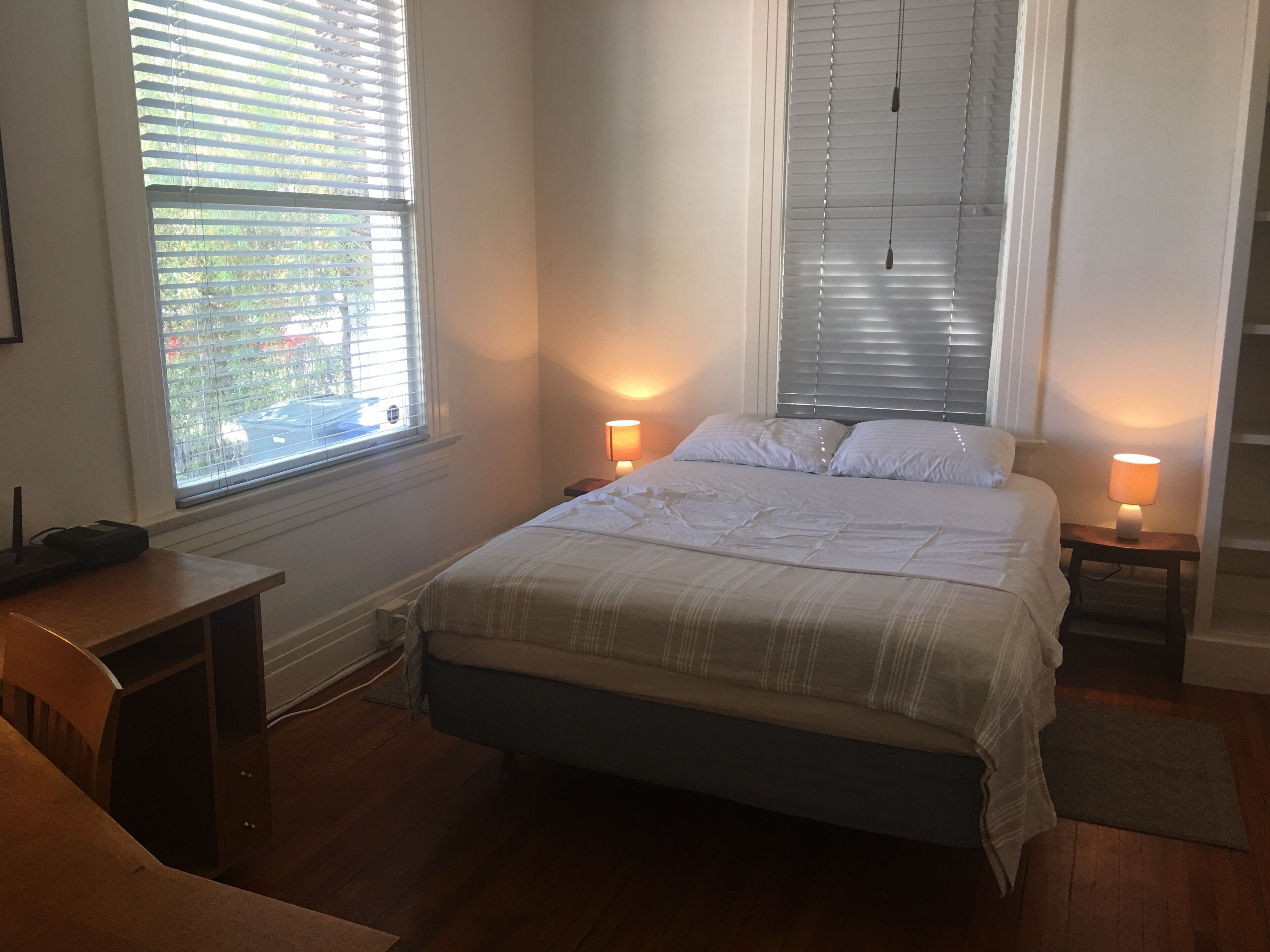 A bedroom with a bed, two bedside tables with lamps, a window with blinds, and a wooden desk.