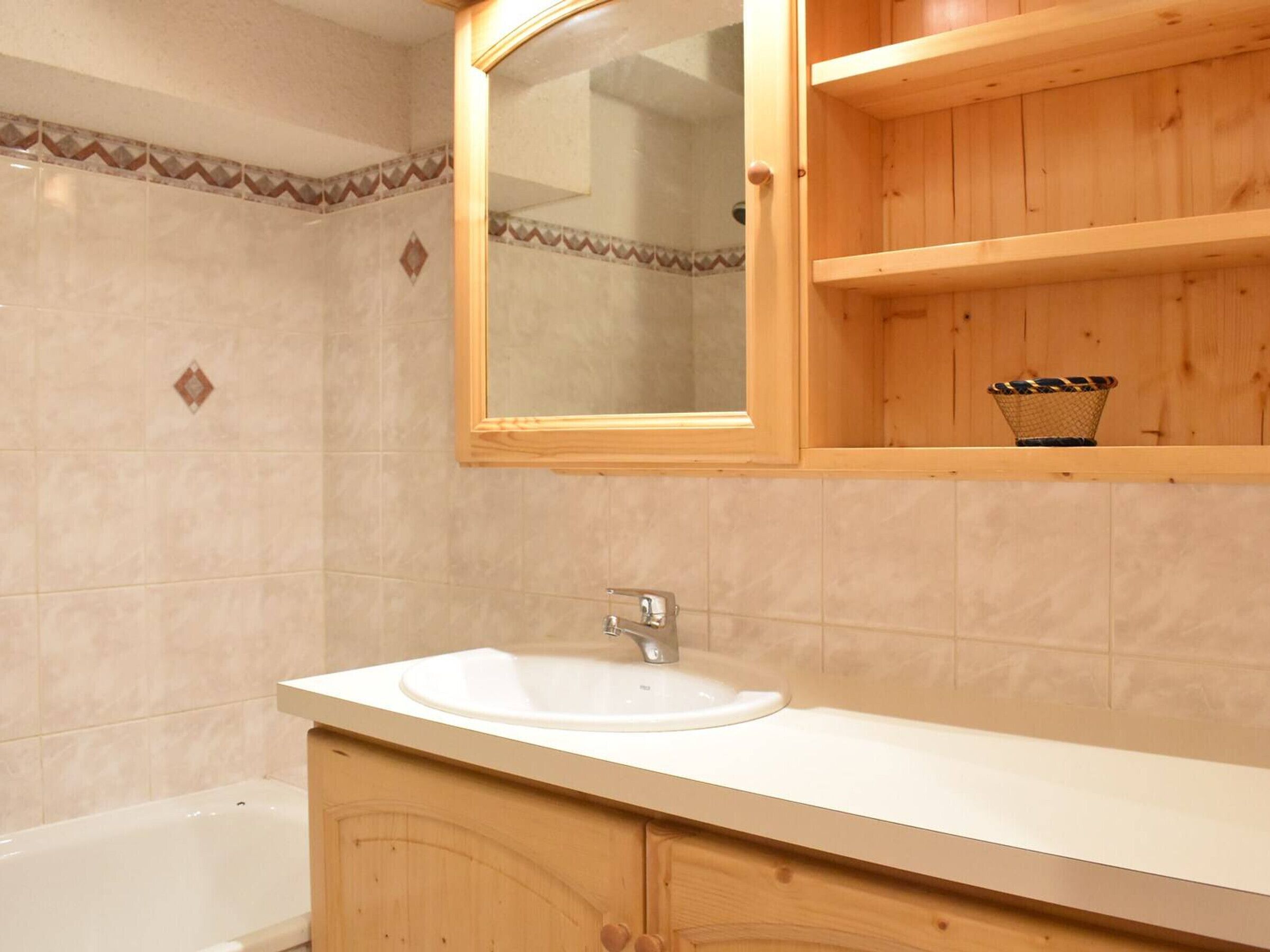 A bathroom with a white sink, wooden cabinets, a mirror, and a small shelf with a basket.