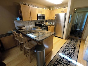 A kitchen with wooden cabinets, stainless steel appliances, a granite countertop, and a patterned rug.