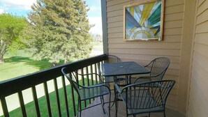 A balcony with a table and chairs, a painting on the wall, and a view of a grassy area with trees. - Private deck, BBQ Grill, view of golf course (Eden)