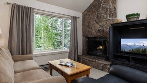 A living room with a stone fireplace, a sofa, a coffee table, and a television displaying a mountain view.