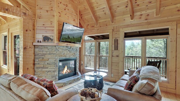 A cozy cabin interior with a stone fireplace, wooden ceiling, and a comfortable seating area with a television mounted above.