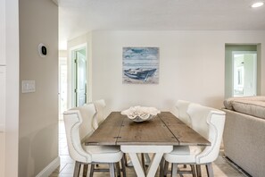 A dining area with a wooden table and white upholstered chairs, a decorative bowl on the table, and a painting of boats on the wall.