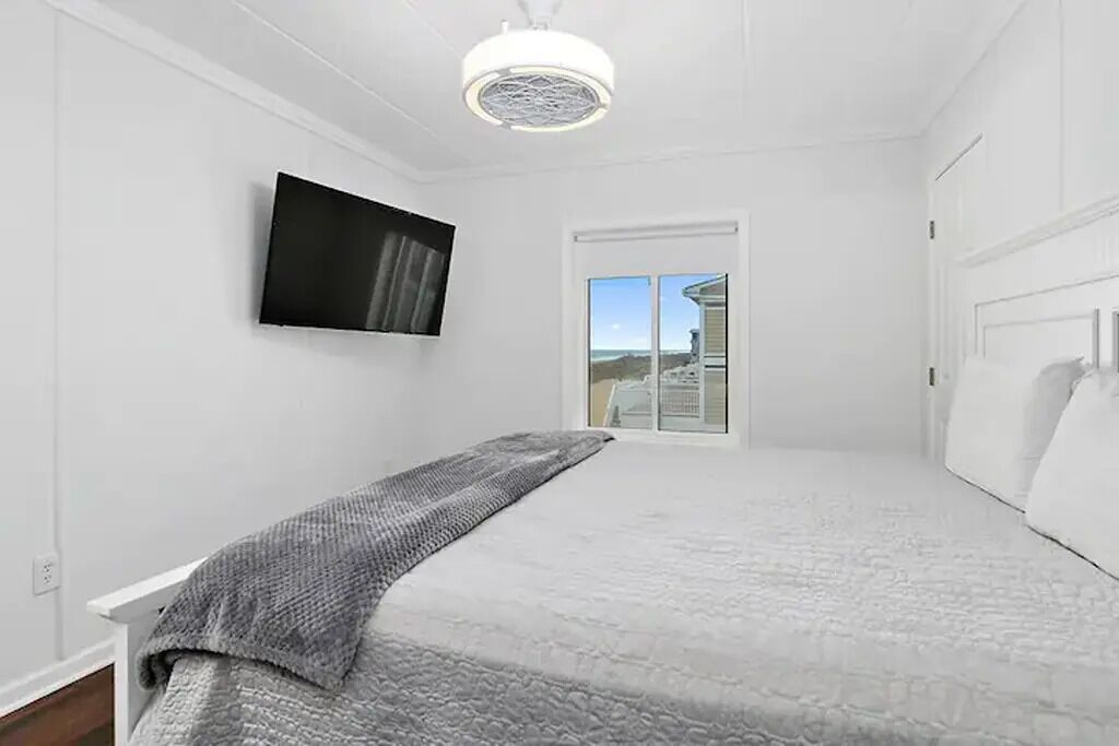 A bedroom with a bed, a flat-screen TV mounted on the wall, a window with a view, and a ceiling light fixture.