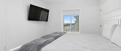 A bedroom with a bed, a flat-screen TV mounted on the wall, a window with a view, and a ceiling light fixture.