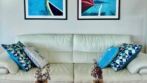 Ein Wohnzimmer mit einem weißen Sofa, zwei gemusterten Kissen, einem Glastisch und zwei gerahmten Bildern vom Meer.