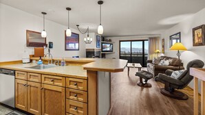 Una cocina moderna con armarios de madera, una isla central e iluminación colgante. Hay un comedor con un sofá y un televisor. La habitación tiene un ventanal con vistas a las montañas.