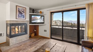 Una sala de estar con chimenea, un televisor montado encima y un amplio ventanal corredizo que da a un balcón con vistas a la montaña.