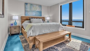 Un dormitorio con cama, mesitas de noche, un banco, un ventanal con vistas y un cuadro en la pared.
