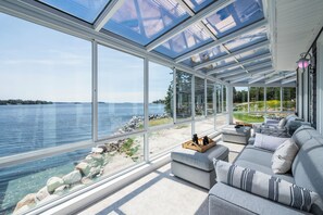 A modern living room with a large glass wall overlooking a lake, a gray sofa, and a wooden coffee table. - The Land Yacht – Indoor Pool, Hot Tub, Solarium Lounge & Sublime Ocean Views (Ingramport)