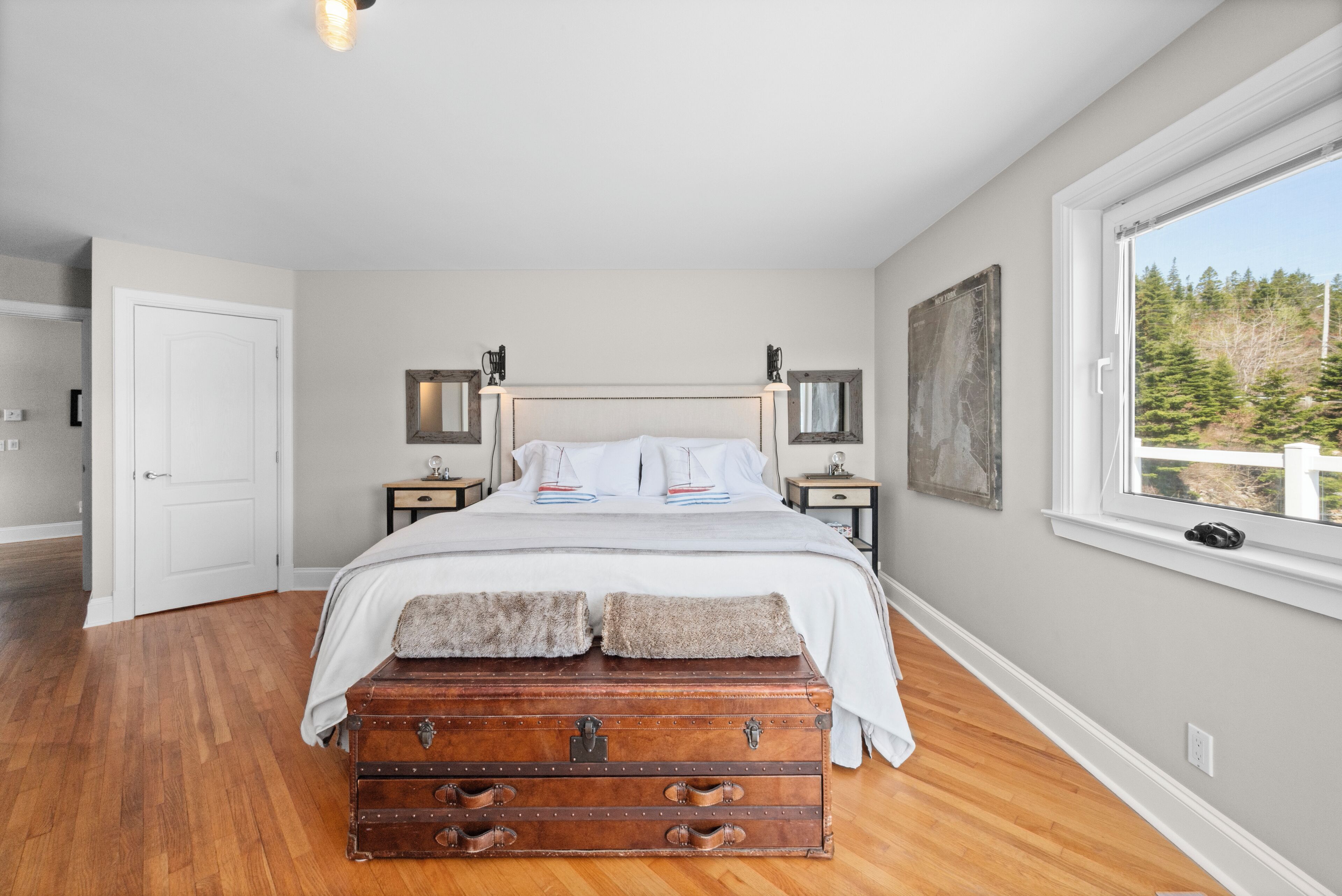 A bedroom with a wooden floor, a bed with white bedding, a vintage trunk, a large window with a view of trees, and a door leading to another room.