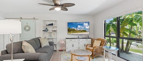 Una sala de estar moderna con un sofá gris, una mesa de centro redonda y una silla de mimbre. Hay una estantería con objetos decorativos y un cuadro de una playa.