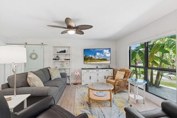 Una sala de estar moderna con un sofá gris, una mesa de centro redonda y una silla de mimbre. Hay una estantería con objetos decorativos y un cuadro de una playa.