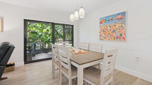 A dining area with a wooden table and chairs, a large window with a view of greenery, and a colorful painting on the wall.