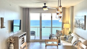A spacious living room with a large TV, a dining table, and a view of the ocean through the sliding glass doors.