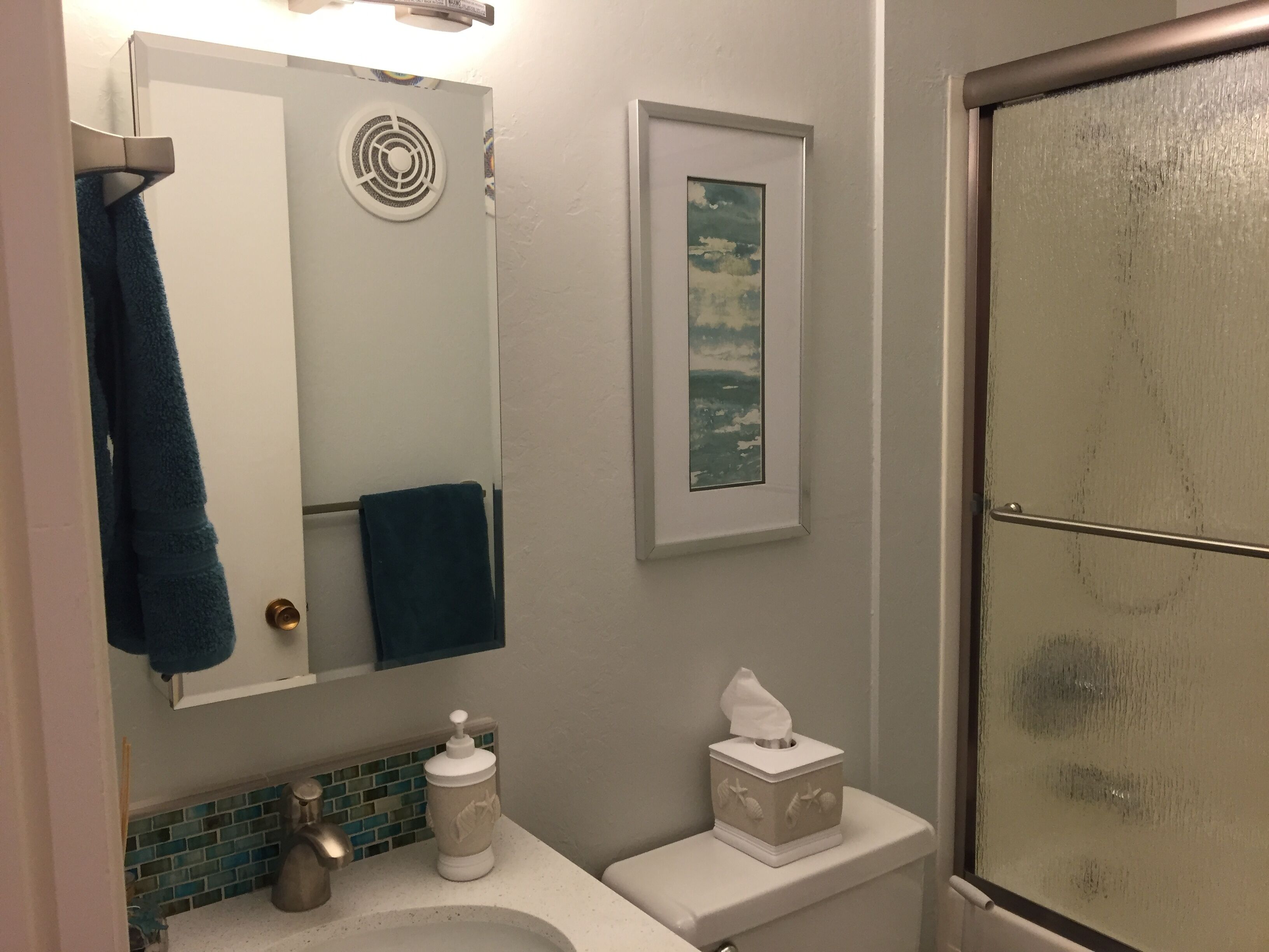 A bathroom with a white sink, a towel rack with a blue towel, a framed picture on the wall, a toilet paper holder, and a shower with a glass door.