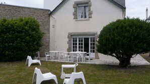 Ein Haus mit Naturstein-Fassade, heller Außenfarbe und einem kleinen Garten mit weißen Kunststoffmöbeln.