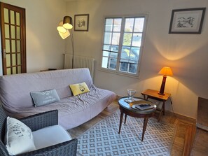 A living room with a sofa, cushions, a small table with a lamp, a window, and a radiator.