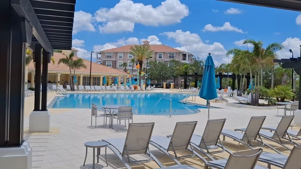 A pool area with lounge chairs and umbrellas, a building in the background, and a clear sky.