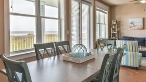 A dining area with a table, chairs, and a decorative glass piece. There are large windows with blinds, a ceiling fan, and a view of the ocean.
