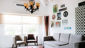 A modern living room with a grey sofa, two armchairs, a coffee table, and a chandelier. There are large windows with blinds, a wall decorated with vintage signs and posters, and an air conditioning unit. - Marina Golf Condo in the heart of the Marina Vallarta (Marina Vallarta)