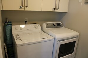 Un espace dédié à la lessive, équipé d’un lave-linge et d’un sèche-linge à chargement frontal, d’une prise électrique et d’un sac à linge bleu suspendu à un étendoir.